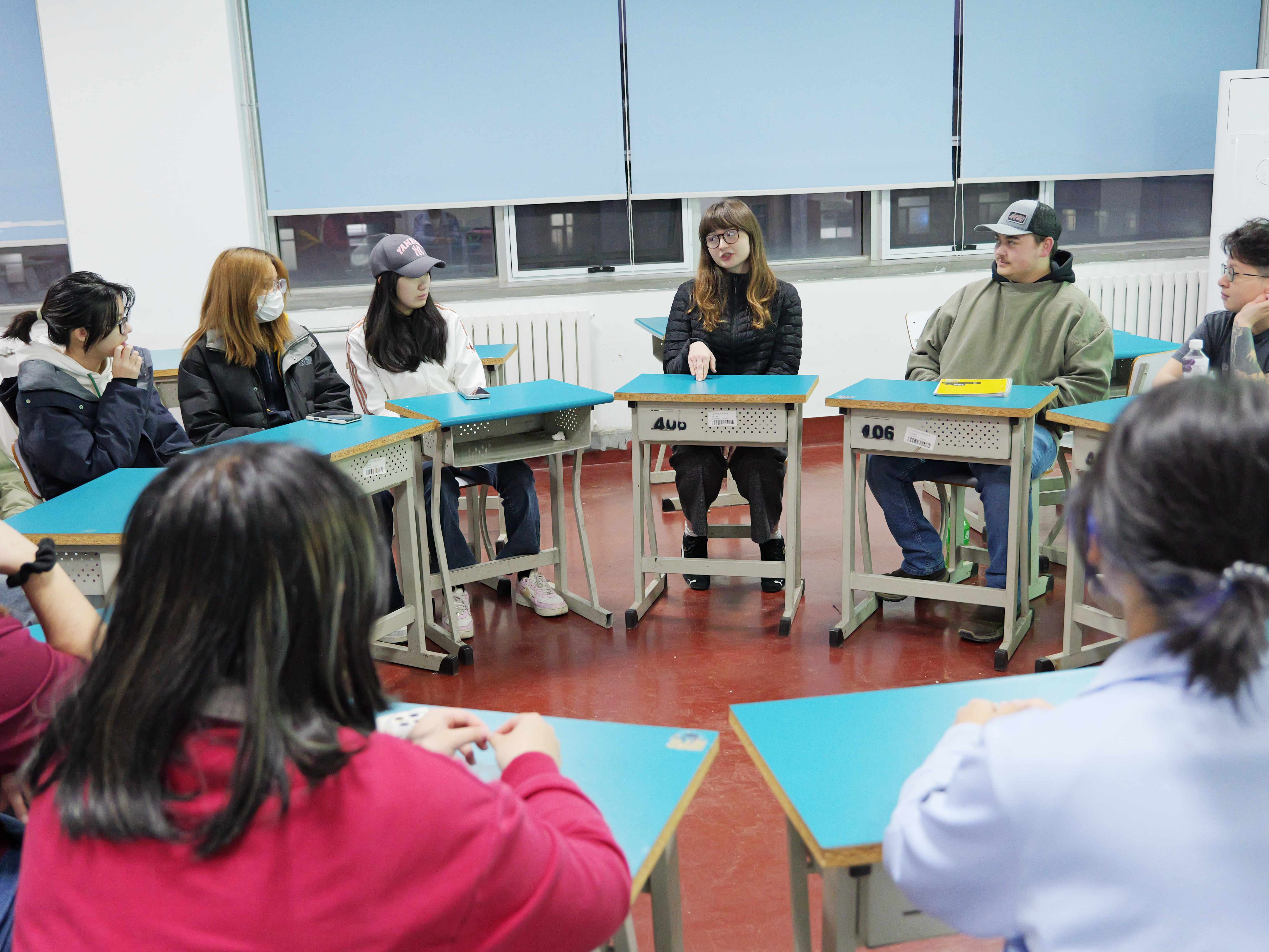 A group of students sitting at desks arranged in a circle inside a classroom, engaged in a discussion.